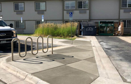 A sidewalk with a bike rack ends abruptly in front of a grassy area, blocking wheelchair access to two accessible parking spots near an apartment building. A dumpster is nearby. A sidewalk with a bike rack ends abruptly in front of a grassy area, blocking wheelchair access to two accessible parking spots near an apartment building. A dumpster is nearby.