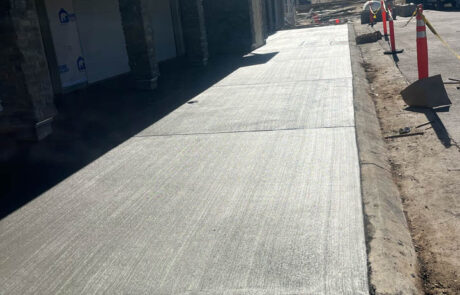 Freshly poured concrete driveway outside a house under construction, with traffic cones, construction equipment, and a modern building visible in the background under a clear blue sky. Freshly poured concrete driveway outside a house under construction, with traffic cones, construction equipment, and a modern building visible in the background under a clear blue sky.