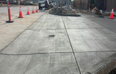 Freshly poured concrete sidewalk curves beside a building under construction. Orange traffic cones line the edge, and construction materials and workers are visible in the background under a cloudy sky. Freshly poured concrete sidewalk curves beside a building under construction. Orange traffic cones line the edge, and construction materials and workers are visible in the background under a cloudy sky.