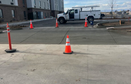A white utility truck is parked on a freshly paved area near a modern multi-story building. Orange traffic cones block off the new pavement under a partly cloudy sky. A white utility truck is parked on a freshly paved area near a modern multi-story building. Orange traffic cones block off the new pavement under a partly cloudy sky.