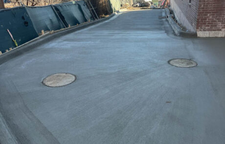 Freshly poured concrete driveway with two round utility covers, bordered by bricks on one side and a fence and trees on the other, with a house and street visible in the background. Freshly poured concrete driveway with two round utility covers, bordered by bricks on one side and a fence and trees on the other, with a house and street visible in the background.