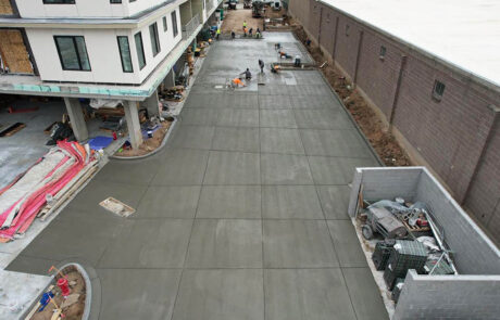 A group of construction workers smooth and finish freshly poured concrete in a large driveway area beside a modern building and a tall brick wall. Construction materials and equipment are scattered nearby. A group of construction workers smooth and finish freshly poured concrete in a large driveway area beside a modern building and a tall brick wall. Construction materials and equipment are scattered nearby.
