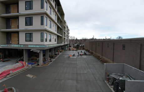 Construction workers are smoothing freshly poured concrete in a wide driveway beside a multi-story building under construction, with materials and equipment scattered around the site. Construction workers are smoothing freshly poured concrete in a wide driveway beside a multi-story building under construction, with materials and equipment scattered around the site.