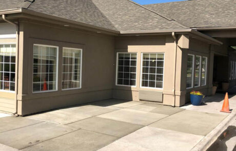 A beige building with multiple large windows, a sloped roof, and a paved sidewalk in front. There is a blue flower pot and orange traffic cones near the entrance. The sky is clear and blue. A beige building with multiple large windows, a sloped roof, and a paved sidewalk in front. There is a blue flower pot and orange traffic cones near the entrance. The sky is clear and blue.