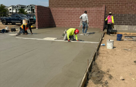 Four construction workers smooth and finish freshly poured concrete on a building foundation at a construction site, with brick walls and nearby buildings visible in the background. Four construction workers smooth and finish freshly poured concrete on a building foundation at a construction site, with brick walls and nearby buildings visible in the background.