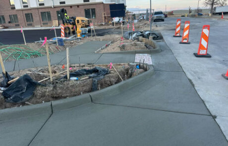 Freshly poured concrete sidewalks and curbs curve around a construction site with soil, stakes, and construction equipment visible. Orange safety barriers and a stop sign are in the background near a street and a building. Freshly poured concrete sidewalks and curbs curve around a construction site with soil, stakes, and construction equipment visible. Orange safety barriers and a stop sign are in the background near a street and a building.