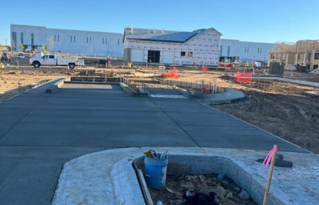 A construction site with freshly poured concrete, a blue bucket and tools in the foreground, and a partially built house with a white exterior in the background under a clear blue sky. A construction site with freshly poured concrete, a blue bucket and tools in the foreground, and a partially built house with a white exterior in the background under a clear blue sky.