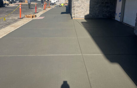 Freshly poured concrete driveway in front of a house under construction, with visible expansion joints. Construction workers and equipment are in the distance, and homes are visible in the background under a clear blue sky. Freshly poured concrete driveway in front of a house under construction, with visible expansion joints. Construction workers and equipment are in the distance, and homes are visible in the background under a clear blue sky.