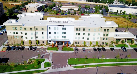 completed-commercial-project-1 Aerial view of a modern, three-story hotel with a white exterior, surrounded by a parking lot with several parked cars, landscaped lawns, and neighboring buildings in the background.