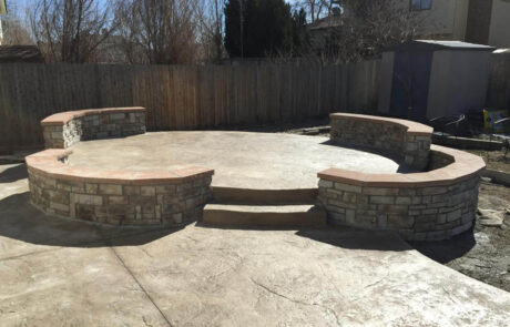 A backyard patio with a circular, stamped concrete surface and two curved stone benches with stone backs and flat tops. There are two steps leading up to the patio. A wooden fence and shed are in the background.