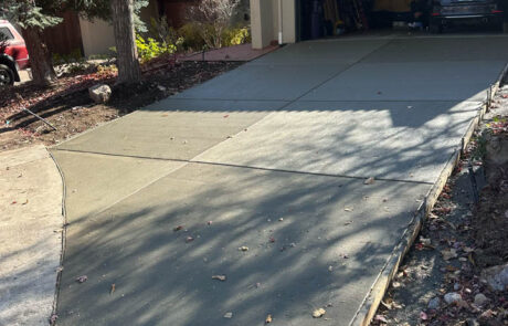 Freshly poured concrete driveway leads to an open garage with a car inside. Shadows of tree branches fall across the driveway. There are trees and landscaping around the house.