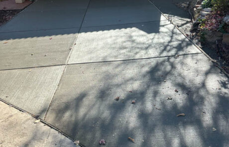 A driveway with freshly poured, smooth concrete leads to an open garage with a car parked inside. Sunlight casts tree shadows on the driveway, and yellow caution tape is visible on the right side.