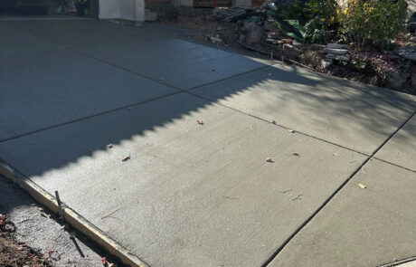 Freshly poured concrete driveway with visible seams, next to a garage with an open door. Yard debris and tools are nearby, and a person is working in the background near trees and landscaping.