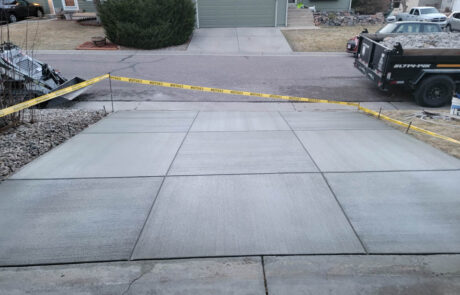 A newly poured concrete driveway sectioned into squares, roped off with yellow caution tape, in front of a house with a green garage door. A black utility trailer is parked nearby.