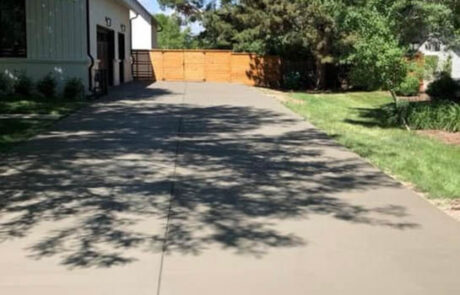A newly paved concrete driveway leads to a building with white siding and a tan wooden fence at the end, surrounded by green grass, trees, and landscaping on a sunny day.