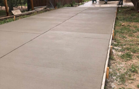 Freshly poured and smoothed concrete driveway with workers in safety vests in the background. The driveway is bordered by wooden forms, with houses, trees, and fencing visible alongside.