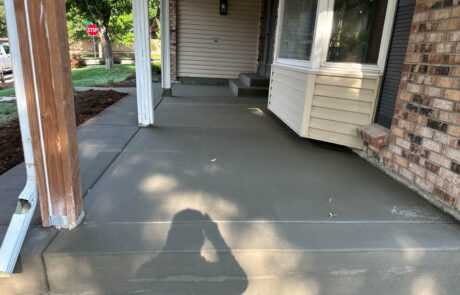 Freshly poured concrete covers a front porch and walkway of a house with brick and siding. A shadow of a person taking the photo is visible on the concrete, and there are a few small leaves on the surface.