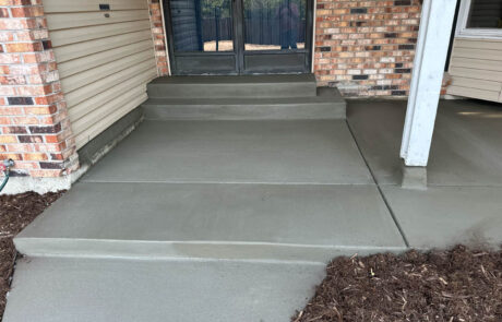 Freshly poured and smoothed concrete steps and walkway lead to a glass door entrance of a brick and siding building; mulch lines the edge of the walkway.