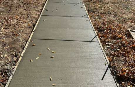 Freshly poured concrete sidewalk with a few scattered leaves on top, surrounded by dirt and some autumn leaves. Safety cones and caution tape are visible in the background near the street.