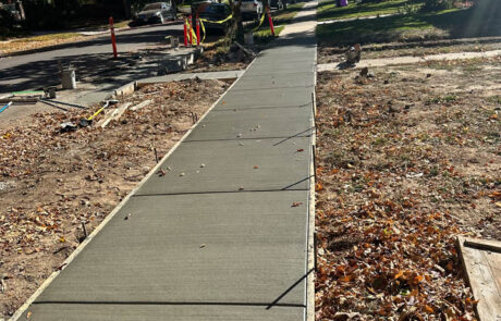 Freshly poured concrete sidewalk runs straight through a leafy neighborhood with autumn foliage, bordered by unfinished construction and scattered fallen leaves. Sunlight filters through the trees, casting shadows on the path.