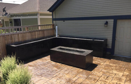 A paved patio with a wooden fence, featuring a rectangular raised planter in the center and an L-shaped black raised planter along the corner near a house with light siding and a white door.