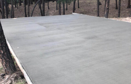 A freshly poured and smoothed concrete slab sits in a forested area surrounded by tall pine trees, with no structures yet built on the slab.